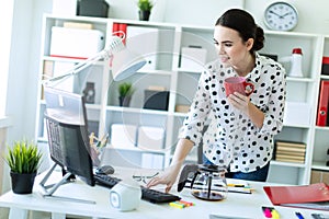 A young girl is standing in the office near the table, holding a red mug in her hand and typing on the keyboard.