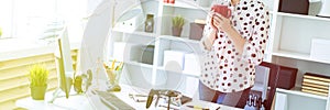 A young girl is standing in the office near the table, holding a red mug in her hands and looking at the monitor.