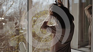 A young girl is standing at the open window closing her eyes and fingering her hair with her hands enjoying the warmth