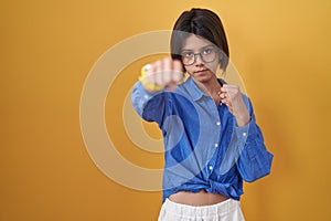 Young girl standing over yellow background punching fist to fight, aggressive and angry attack, threat and violence