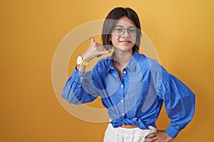 Young girl standing over yellow background smiling doing phone gesture with hand and fingers like talking on the telephone