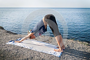 Young girl is standing in yoga dog pose on a stony sea coast in summer morning.