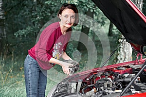A young girl stands at a broken car and holds a bad spare part, an electric generator, does not understand how to repair