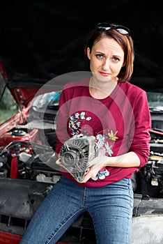 A young girl stands at a broken car and holds a bad spare part, an electric generator, does not understand how to repair