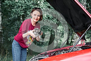 A young girl stands at a broken car and holds a bad spare part, an electric generator, does not understand how to repair