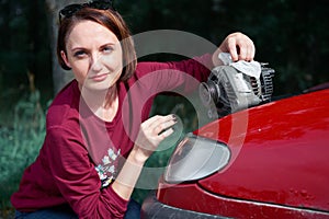 A young girl stands at a broken car and holds a bad spare part, an electric generator, does not understand how to repair, clean it