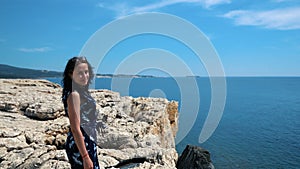 Young girl stands on the edge of a cliff and looks at the sea. Beautiful girl enjoying a view at the cliff above the sea