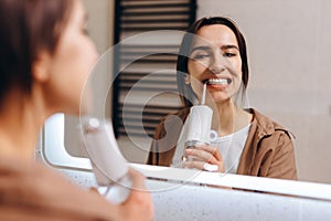 A young girl stands in front of a mirror in the bathroom and rinses her mouth with an irrigator photo