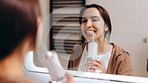 A young girl stands in front of a mirror in the bathroom and rinses her mouth with an irrigator photo