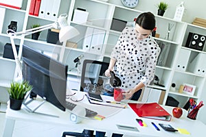 A young girl stands in the office near the table and pours coffee from the coffee pot into a red cup.