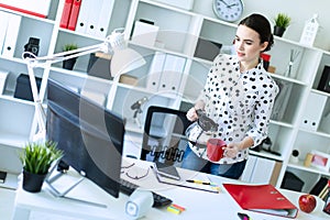 A young girl stands in the office near the table and pours coffee from the coffee pot into a red cup.