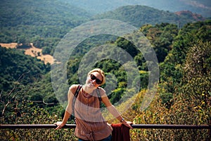 Young girl stands on viewpoint and smiles on sunny day. Happy female tourist enjoys beautiful panorama. Cute stylish girl in