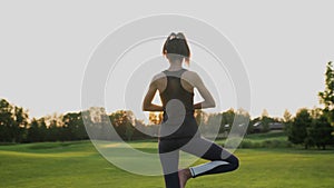 A young girl stands in a warrior pose on green grass doing yoga at sunset time