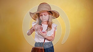 Young girl in straw hat holding lipstick and paints lips with lipstick. Smiling child with lipstick and mirror in hand.