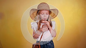 Young girl in straw hat holding lipstick and paints lips with lipstick. Smiling child with lipstick and mirror in hand.