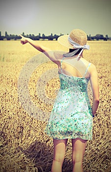 young girl with straw hat in the wheat field  pointing to a nearby place with the index finger and antiqued toned effect