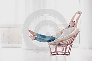 Young girl is stretching in modern armchair in light livingroom. She is sleepy and dreamy, wearing casual clothes