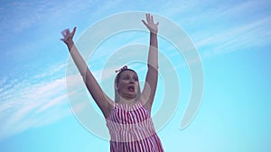 Young girl in a striped red and white dress. A girl stands against a blue sky, raising her hands up, a sign of freedom.