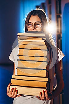 Young girl student with glasses in library smiling and carries stack of books. Exam preparation