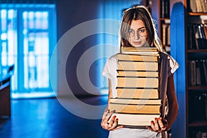 Young girl student with glasses in library smiling and carries stack of books. Exam preparation