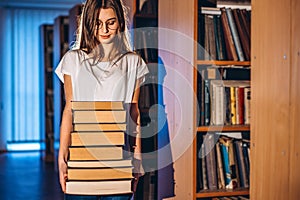 Young girl student with glasses in library smiling and carries stack of books. Exam preparation