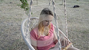 A young girl student reading a book on a swing in the park at sunset. She straightens her hair and swings on a white