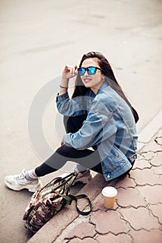 Young girl student is sitting on the pavement. Wear jeans and drink coffee. Young beautiful model girl in sunglasses smiles