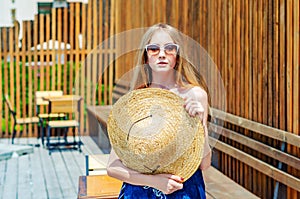 Young girl in sunglasses holds a big straw hat in her hands, shields her face