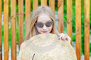 Young girl in sunglasses holds a big straw hat in her hands, shields her face