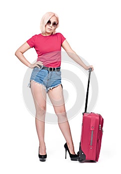 A young girl in sunglasses, a pink T-shirt and denim shorts stands with a pink suitcase, isolated on white