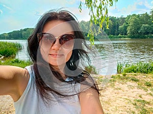 Young girl in sunglasses in white T-shirt smiling on river bank. Young smiling woman tourist standing, wearing white