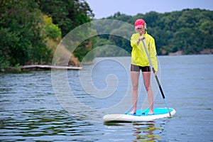 Young girl-surfer riding on the stand-up paddle board in the clear waters of the on the background of green trees