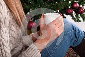 Young Girl in sweater holding cup of coffee in hand at legs with warmers and Christmas tree in the background