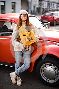 Young girl in sweater and jeans stands near a red vintage car on a street with a flowers