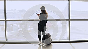 Young girl takes selfie photo near airport window. Happy European tourist with backpack uses smartphone in terminal. 4K.
