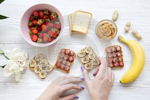 Young girl takes vegan toast with fruits, seeds, peanut butter over white wooden background, top view. Healthy breakfast with ingr