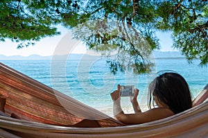 Young girl taking a picture with a smartphone lying in a hammock on a beach