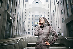 Young girl talking on mobile phone in courtyard business center. girl with long dark hair dressed in winter jacket in cold weather