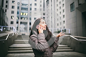 Young girl talking on mobile phone in courtyard business center. girl with long dark hair dressed in winter jacket in cold weather