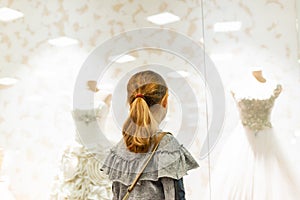 A young girl, a teenager, stands near the window of a wedding salon and looks at wedding dresses, introducing family life