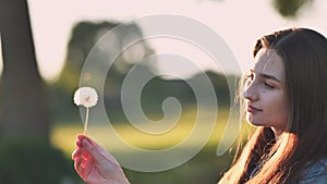 A young girl tenderly looks at a dandelion flower on a summer evening.