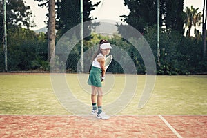 Young girl, tennis player with green skirt, white top and hat, playing tennis on a clay tennis court