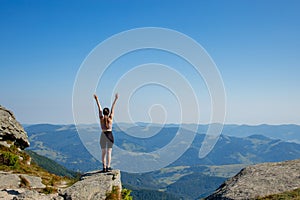 The young girl at the top of the mountain raised her hands up on blue sky background. The woman climbed to the top and enjoyed her