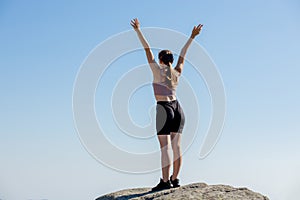 The young girl at the top of the mountain raised her hands up on blue sky background. The woman climbed to the top and enjoyed her