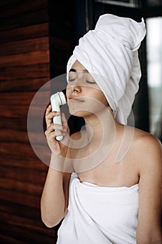 Young girl with towel on her head makes facial massage with wireless electric massager