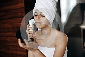 Young girl with towel on her head makes facial massage with wireless electric massager