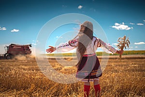 Young girl with traditional Bulgarian folklore costume at the agricultural wheat field during harvest time with industrial combine