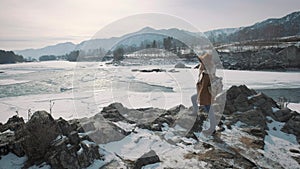 A young girl traveler approaches the edge of the cliff shore. He looks out into the distance to the frozen river in