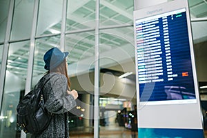 Young girl traveler with a backpack in a hat looks at the information board at the airport. Getting information about