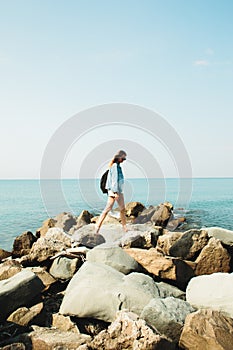 A young girl traveler with a backpack walks barefoot on large stones on the sea coast